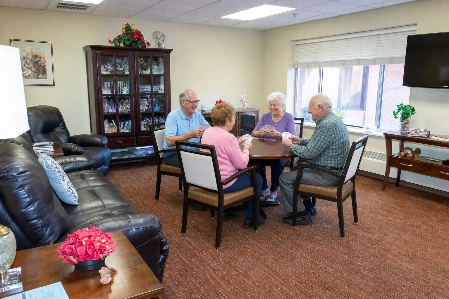 Two men and two women playing cards at a circular table in a living room area with couch, TV, and bookshelf.